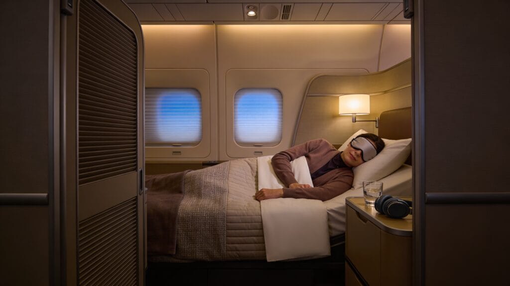 Modern long-haul business class suite with its sliding door fully closed, showing a wide lie-flat seat made into a bed with white bedding and a blanket, a passenger lying on their side asleep with an eye mask on, soft warm ambient lighting, a window shade mostly closed, and a side table holding a glass of water and headphones.